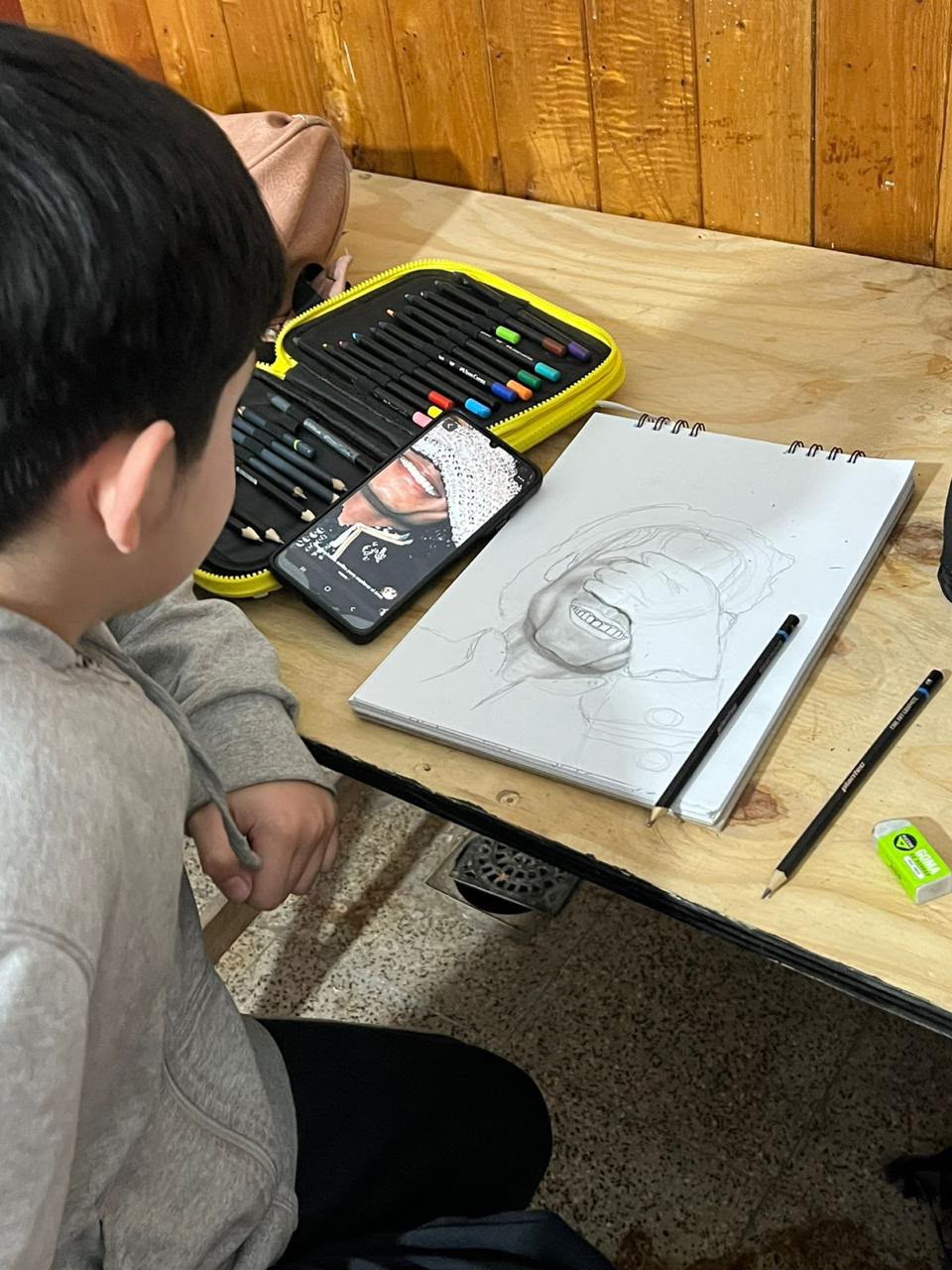 Niño dibujando, visto de espaldas, junto a sus lápices